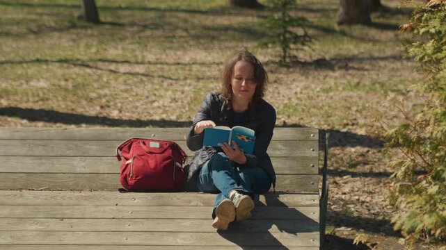 Woman reads book on wooden bench in park with red bag beside her. Book has colorful design woman wears dark jacket and jeans. Sunlight creates shadows around bench and subject