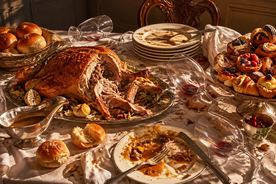 Wide still-life of overloaded banquet table after long meal, torn roast, stacked plates, scattered bread, tipped wine glasses and pastries, gluttony mood