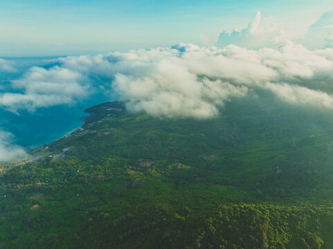 An aerial drone view looking down through thick, soft low clouds and mist rolling over a lush green coastal mountain ridge and ocean shoreline.