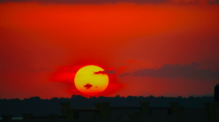 A striking sunset view featuring a vivid glowing red and orange sun dipping below dark cloud layers over a distant horizon. © Rama
