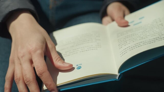 Close up of woman hands turning pages of open book with text and diagrams resting on blue fabric. scene shows consistent soft lighting highlighting hand details and page content