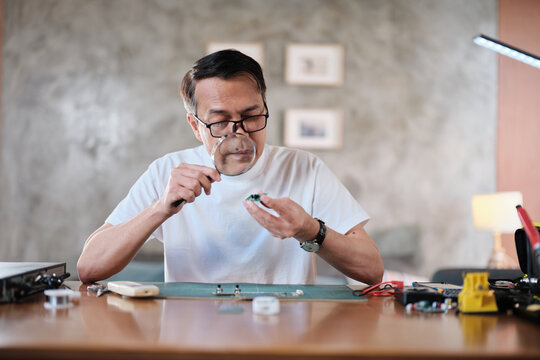 Mature Asian man looks at and considers circuit board details with magnifying glass, focusing on precision checks for home electrical appliance DIY fix, electronics component hobby, handyman workshop.