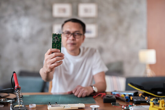 Selective focus on circuit board showing a mature Asian man proud of a successful soldering job, cheerful job achievement, home electronics appliance repair handyman, DIY fix tools, hobby workshop.