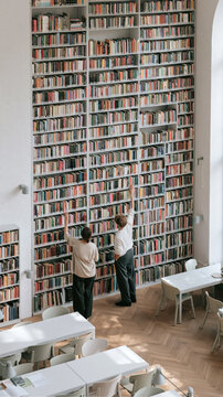 A Journey Through Books: Two individuals reach for knowledge among towering shelves filled with a vast collection of books in a grand library.