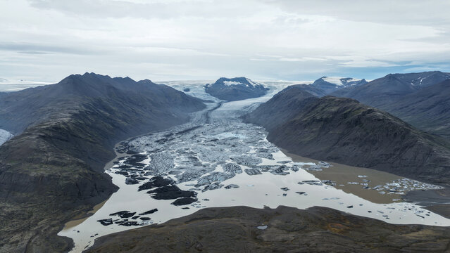 Aerial view of Flaajokull glacier tongue flowing into a glacial lake with icebergs, framed by mountains under an overcast sky in Hofn, Sveitarfelagid Hornafjardarbaer, Iceland.