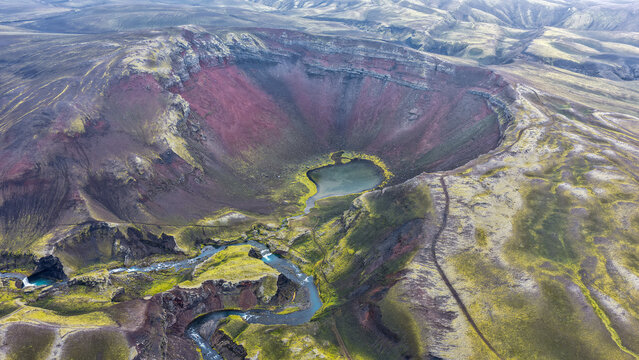 Aerial view of the Raudibotn volcanic crater featuring vibrant red slopes, a central lake, and a winding river cutting through the mossy landscape of Svartafellstangi, Skaftarhreppur, Iceland.