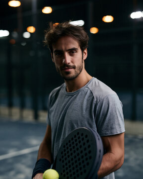 Tennis player holds paddle with focus. Dark room lights highlight his intense gaze. He grips the ball, ready to strike. Athletic stance suggests imminent action. Ideal for sports, fitness