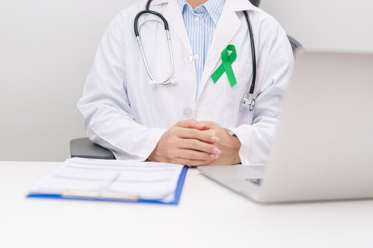 doctor using laptop and report notes on notebook. green ribbon for July Eye Injury Prevention month, January Glaucoma Awareness month, February Gallbladder and Bile duct and October Liver cancer