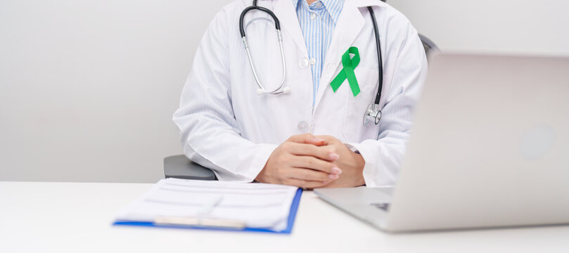 doctor using laptop and report notes on notebook. green ribbon for July Eye Injury Prevention month, January Glaucoma Awareness month, February Gallbladder and Bile duct and October Liver cancer