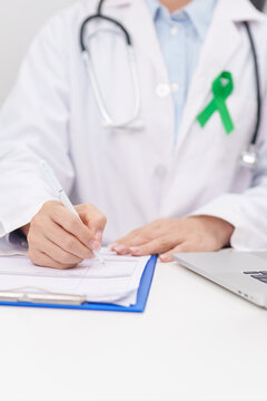 doctor using laptop and report notes on notebook. green ribbon for July Eye Injury Prevention month, January Glaucoma Awareness month, February Gallbladder and Bile duct and October Liver cancer