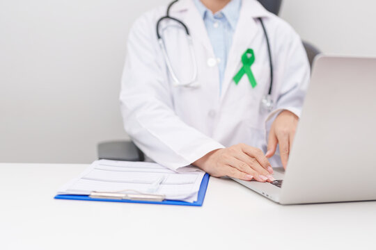 doctor using laptop and report notes on notebook. green ribbon for July Eye Injury Prevention month, January Glaucoma Awareness month, February Gallbladder and Bile duct and October Liver cancer