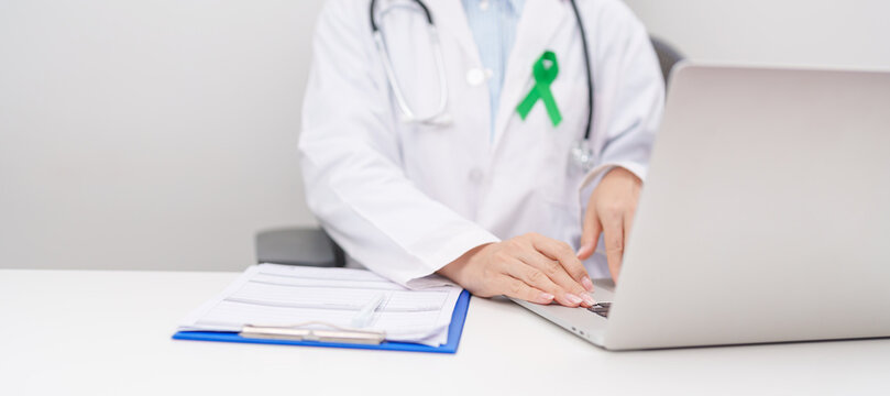doctor using laptop and report notes on notebook. green ribbon for July Eye Injury Prevention month, January Glaucoma Awareness month, February Gallbladder and Bile duct and October Liver cancer