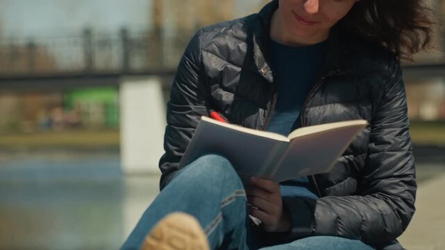 Close up of woman writing in open notebook with red pen while seated outdoors on sunny day. Notebook pages visible as woman writes wearing dark puffer jacket and blue shirt. Background shows
