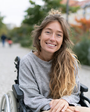 Young woman smiles warmly from wheelchair. Long, wavy hair frames her joyful expression. She sits calmly on a paved path, surrounded by greenery. Background shows blurred trees