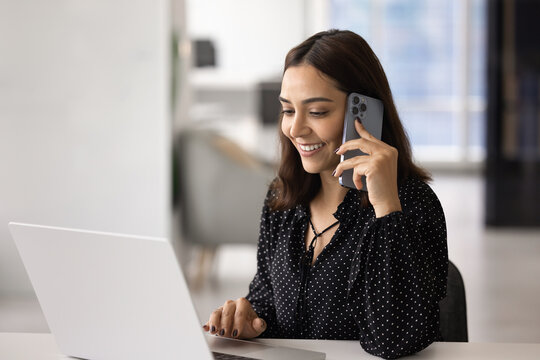 Happy female employee having pleasant phone conversation while working on laptop in contemporary office, engaged in positive, personal or business communication. Remote work, modern technology usage