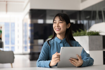 Smiling Asian professional woman looks away thoughtfully, holding tablet while seated at desk in...