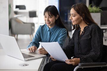 Young Middle-Eastern sits in wheelchair listening female colleague explain corporate software,...