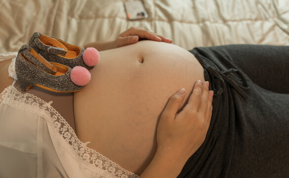 Overhead close-up of a pregnant belly with tiny glittery baby shoes resting on top and both hands cradling it.