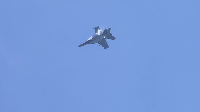 A fighter jet performs an inverted pass with afterburners on and vapor trails forming in a clear blue sky.