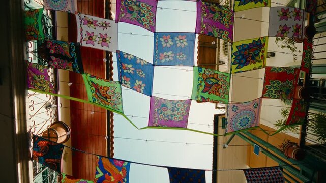Close-up patchwork squares hang indoors. Vibrant floral and geometric patterns appear. Colors range from red to blue and green. Broader view shows quilt hanging indoors