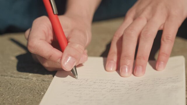 Close up of hands writing with red pen on lined paper outdoors. person writes legibly revealing handwritten text that progresses steadily across page. Soft natural light creates clear