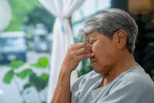 Elderly Asian woman sitting on a sofa at home, covering her eyes due to eye strain and fatigue from prolonged screen use.