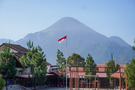 Indonesian flag waving in front of Mount Penanggungan, with pine trees, tiled rooftops, blue sky, and a quiet courtyard forming a scenic highland setting in East Java.