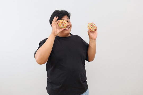 An overweight Asian man stands against a plain white background, posing while holding two doughnuts.