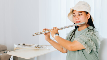 A female flutist in a green shirt sits on a brown couch in her cozy living room, playing a silver flute with concentration © sorapop