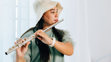 A female flutist in a green shirt sits on a brown couch in her cozy living room, playing a silver flute with concentration © sorapop