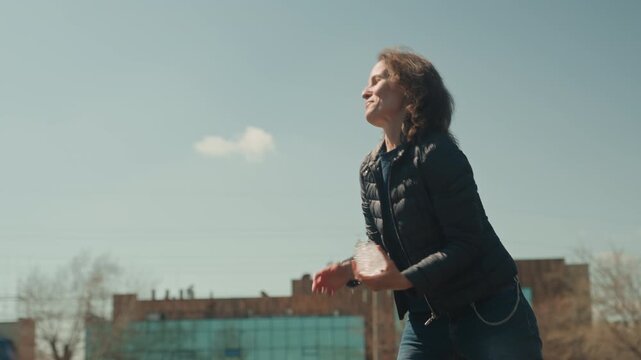 Woman holds and examines block of ice outdoors on sunny day then throws it into air. Woman wears casual clothing including jeans with chain accessory against urban backdrop with buildings