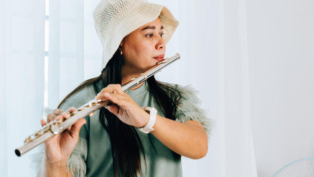 A female flutist in a green shirt sits on a brown couch in her cozy living room, playing a silver flute with concentration