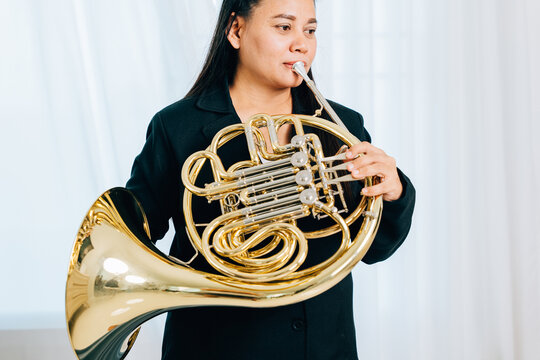 Dressed in black, a woman blows a gleaming French horn in gold