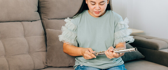 A female flutist in a green shirt sits on a gray sofa in her living room, carefully assembling her silver flute. The scene captures a calm and focused moment as she prepares for her musical session © sorapop