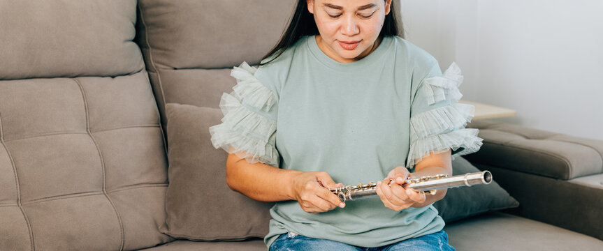 A female flutist in a green shirt sits on a gray sofa in her living room, carefully assembling her silver flute. The scene captures a calm and focused moment as she prepares for her musical session