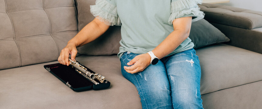A female flutist in a green shirt sits on a gray sofa in her living room, carefully assembling her silver flute. The scene captures a calm and focused moment as she prepares for her musical session