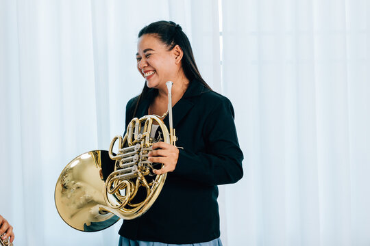 A woman stands at home in front of elegant curtains, playing a shiny gold horn. The warm lighting and her graceful demeanor bring out the beauty of her musical