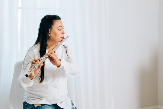 A flutist in a white shirt sits on a chair at home, blowing into her silver flute. The living room curtain backdrop accentuates