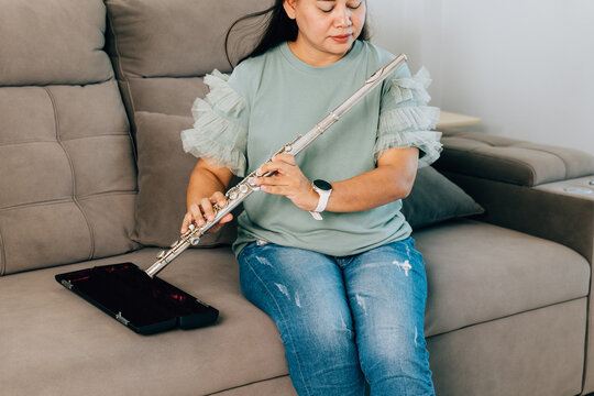 A female flutist in a green shirt sits on a gray sofa in her living room, carefully assembling her silver flute. The scene captures a calm and focused moment as she prepares for her musical session