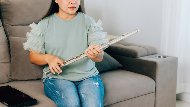 A female flutist in a green shirt sits on a gray sofa in her living room, carefully assembling her silver flute. The scene captures a calm and focused moment as she prepares for her musical session