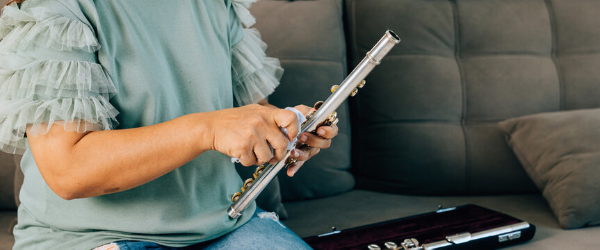 A female flutist sits on a brown couch at home, carefully wiping her silver flute with a soft cloth. The living room setting reflects her dedication to maintaining