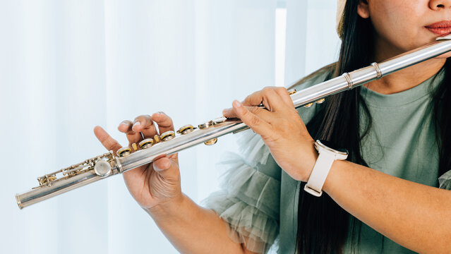 A female flutist in a green shirt sits on a brown couch in her cozy living room, playing a silver flute with concentration