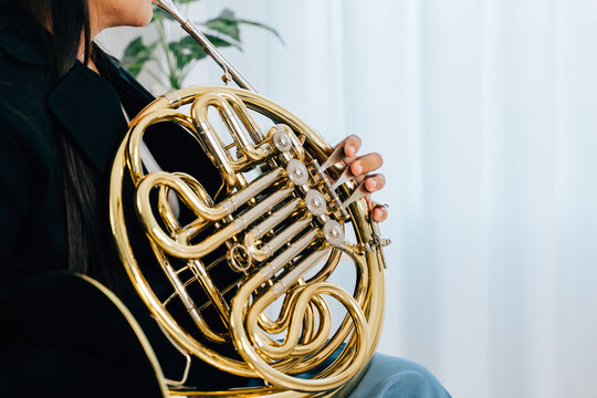A woman sits on a cozy sofa in her living room, playing a shiny gold French horn. The warm lighting and serene atmosphere reflect her passion for music