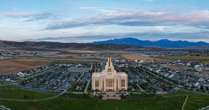 Saratoga Springs, Utah, United States - 22 August 2026: Aerial view of the Saratoga Springs Utah Temple glowing during the blue hour, surrounded by suburban neighborhoods and mountains.