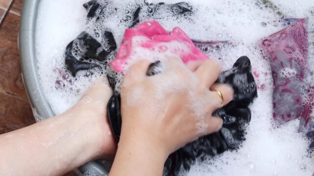 Top view of an Asian woman's hands washing clothes manually in a plastic basin filled with soapy water and foam, doing laundry at home.