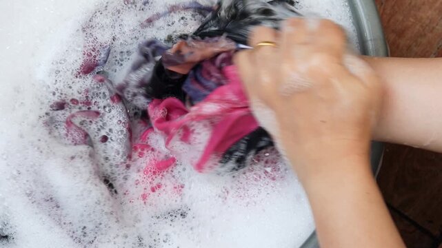 Top view of an Asian woman's hands washing clothes manually in a plastic basin filled with soapy water and foam, doing laundry at home.