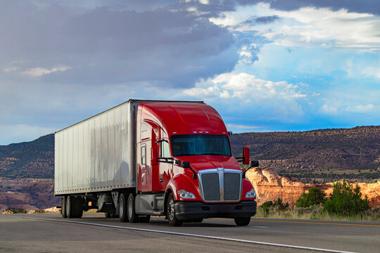 Red semi truck driving on highway through scenic canyon desert landscape with dramatic cloudy sky in USA