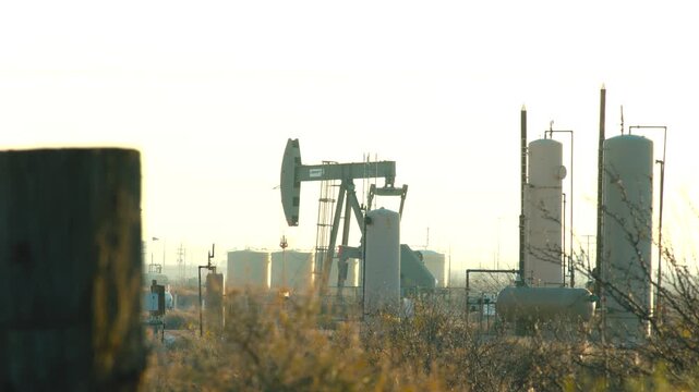 Oil pump jack operating at dusk in west Texas oilfield with storage tanks.