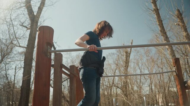 Close up of woman performing pull ups on outdoor exercise bar in daylight wearing casual clothing with sunlight illuminating scene and bare trees visible in background