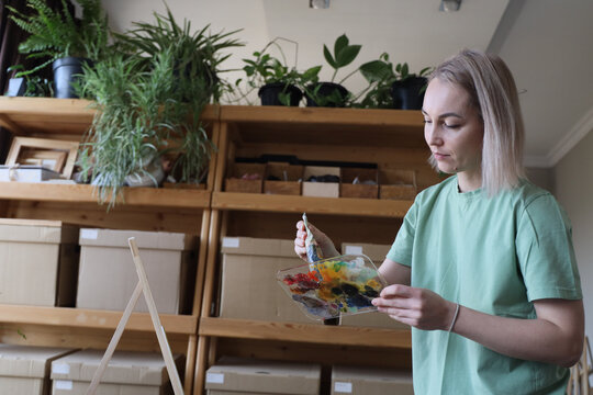 Young Woman In Mint Shirt Studies Colorful Paint Palette While Working In Plant Filled Studio, Surrounded By Wooden Shelves, Potted Greenery, Cardboard Boxes, And Soft Daylight